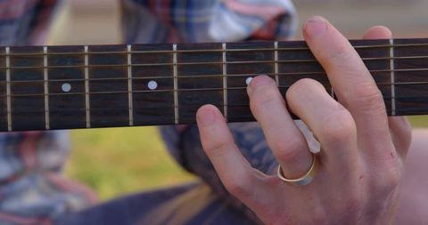 Pressing strings on acoustic guitar fretboard outdoors with plaid shirt and silver ring