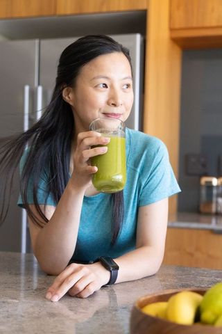 Mature asian woman sipping green smoothie while leaning on kitchen counter wearing black smartwatch