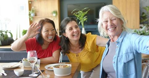 Diverse multi-generational women laughing and posing for selfie at sunlit dining table