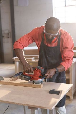 Woodworker smoothing oak plank with electric sander under sunlit workshop window