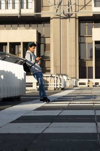 African american teen leaning on rooftop railing checking smartphone with backpack urban