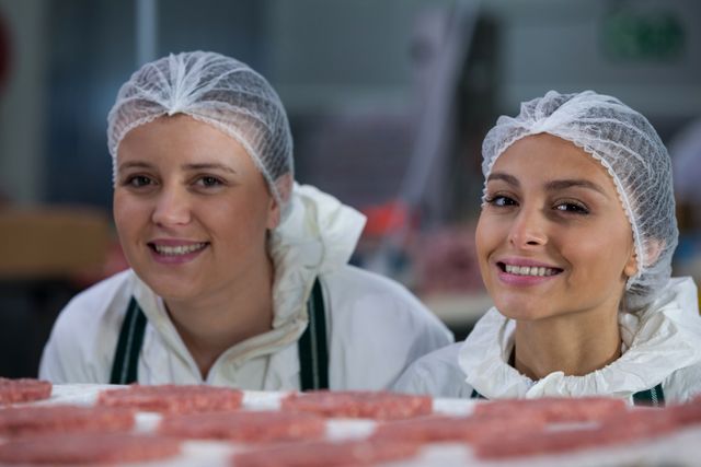 Two female butchers are smiling while working in a meat factory. They are wearing protective clothing and hairnets, ensuring hygiene and safety in the food processing environment. This image can be used for articles or advertisements related to the food industry, meat processing, workplace safety, and professional teamwork.