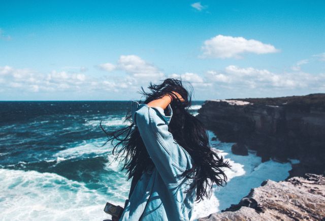 Woman with long hair standing near edge of rocky coastline on breezy day. Ocean waves crashing against cliffs with blue sky and clouds in background. Perfect for travel blogs, adventure-themed projects, scenic nature promotions, or lifestyle imagery.