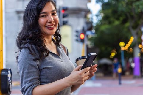African american woman smiling, holding smartphone and coffee on downtown evening street