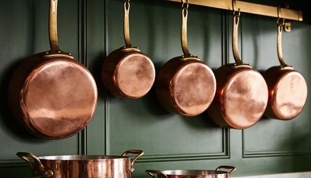 Hanging copper pans on green cabinetry with brass rail and gleaming stockpots for rustic kitchen