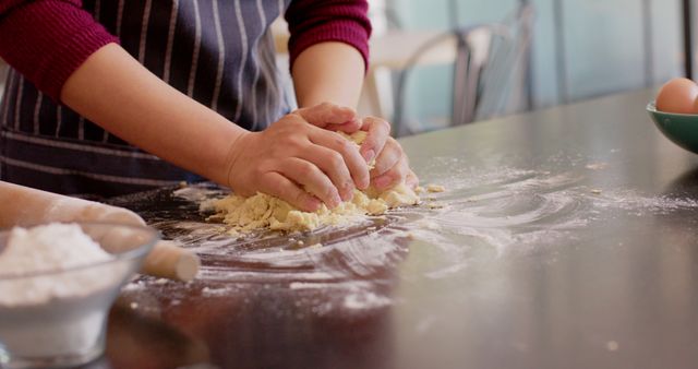 Close-up of person kneading dough on floured kitchen counter. This scene captures the process of making baked goods, perfect for illustrating baking, domestic cooking, food blogs, recipe books, or culinary websites. The raw dough and casual attire add a homely, rustic feel.