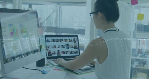 Woman editing media gallery on laptop in modern open-plan office with large monitor in daylight