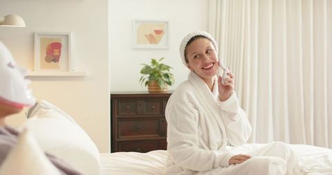 Diverse couple relaxing in robes on bed, woman using facial cleanser, partner wearing led mask