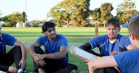 Male cricket teammates sitting on field chatting and planning strategy during outdoor practice