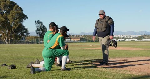 Umpire walking to mound holding mask while diverse baseball players kneeling in green uniforms