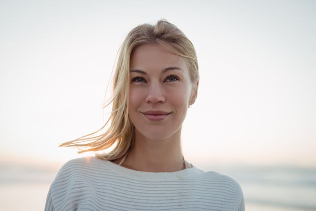 Young woman with blonde hair standing at the beach during dusk, looking away thoughtfully. Ideal for use in lifestyle blogs, mental health articles, travel websites, and advertisements promoting relaxation and mindfulness.