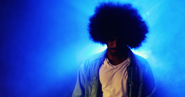 Man with afro hairstyle standing in atmospheric blue misty lighting wearing denim jacket and white t-shirt. Bright backlighting creates dramatic, moody look with slight silhouette effect. Ideal for use in advertisements, fashion editorials, music related themes, or artistic posters.