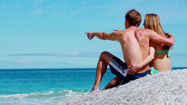 A couple sitting on a sandy beach near ocean, hugging each other warmly while looking at horizon. Great for travel and tourism promotions, romantic getaway advertisements, vacation brochures, summer love themes, and peaceful beach lifestyle content.