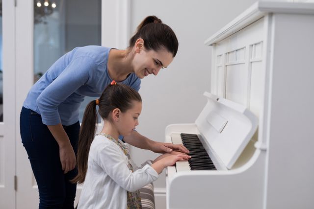 This image captures a mother teaching her young daughter to play the piano at home. It is perfect for use in educational materials, parenting blogs, music lesson advertisements, and family-oriented content. The scene highlights the importance of parental involvement in children's learning and the joy of shared activities.