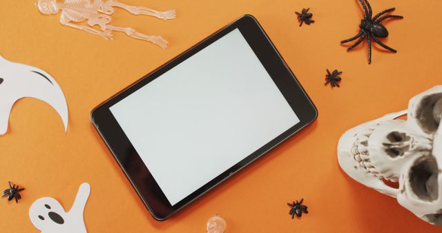 Digital tablet surrounded by Halloween-themed decorations like skeletons, spiders, ghosts, and a skull on an orange background. Suitable for designs related to Halloween technology, digital Halloween celebrations, educational Halloween lessons, or festive autumn themes.