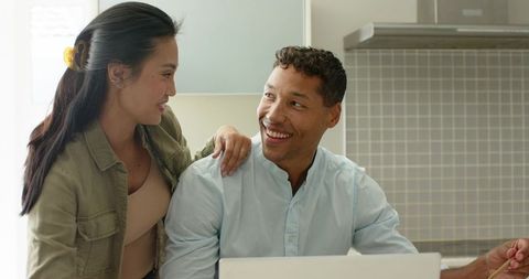 Diverse couple collaborating on laptop in bright modern kitchen, smiling together