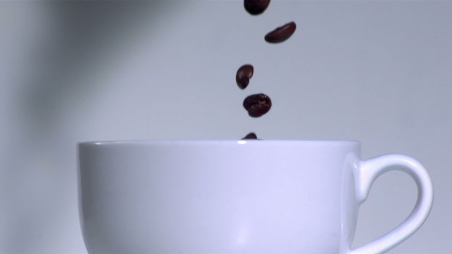 Picture captures fresh coffee beans falling gracefully into a white cup, with gentle steam providing a backdrop. Perfect for coffee shop decorations, ads for coffee ingredients, or blog posts focused on coffee culture and the brewing process. This image conveys freshness, elegance, and a sense of mesmerizing calm associated with preparing a fresh cup of coffee.