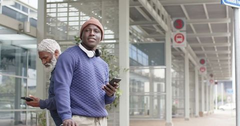 Young African American Commuter Waiting with Smartphone and Headphones under Urban Transit Canopy
