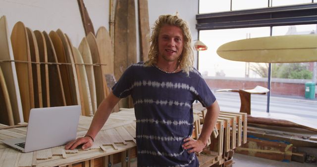 Young male surfer standing in a surfboard workshop, leaning on a wooden table with a laptop. He has a confident and relaxed demeanor, wearing casual attire. Various surfboards and woodworking tools are visible in the background, creating a coastal, creative atmosphere. Ideal for depicting small business entrepreneurship, surf culture, creative crafting industries, and remote workspaces.