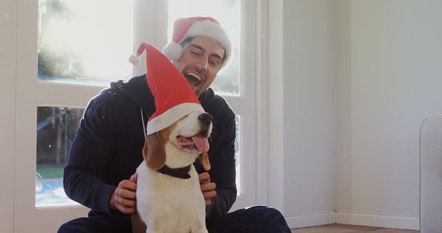 A young Caucasian man wearing a Santa hat shares a joyful moment with his beagle, also sporting a festive hat, with copy space. Their laughter and holiday attire suggest a cheerful celebration of the Christmas season.