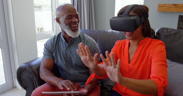 Senior African American couple enjoying virtual reality experience at home together. Man holding tablet while woman is using VR headset, interacting with virtual environment. Perfect for illustrating senior engagement with new technology, modern lifestyle, leisure activities for the elderly, and technology in everyday life.