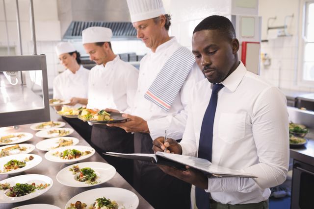 Restaurant manager supervising kitchen staff in a commercial kitchen. Chefs in uniform preparing dishes while manager takes notes. Ideal for illustrating concepts of restaurant management, teamwork in the culinary industry, and professional kitchen environments.