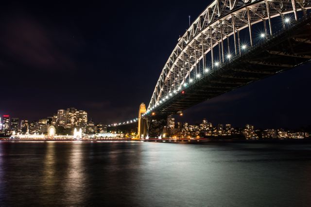 Sydney Harbour Bridge illuminated at night with city skyline as a backdrop. Bright lights from the bridge and city reflecting on the water add to the lively atmosphere. This image is perfect for use in travel promotions, city guides, or articles highlighting urban beauty and famous landmarks.