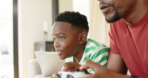 African american dad and son playing video games on sofa, family bonding at home