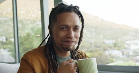 Middle-aged man with dreadlocks sipping from green mug by window overlooking hillside