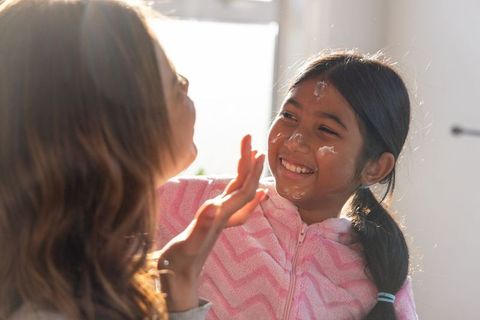Mother applying cream to asian daughter wearing pink fleece in sunlit morning