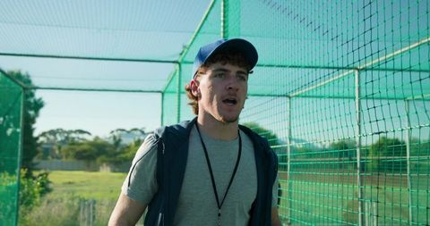 Coach coaching inside netted cage wearing cap and whistle during outdoor baseball training