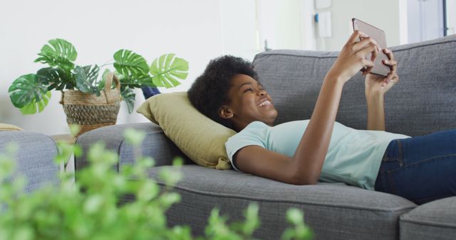 Young woman lying on a gray couch, smiling while using a tablet. Indoor setting with houseplant on side. Suitable for themes like relaxation, technology use, digital lifestyle, and home comfort.