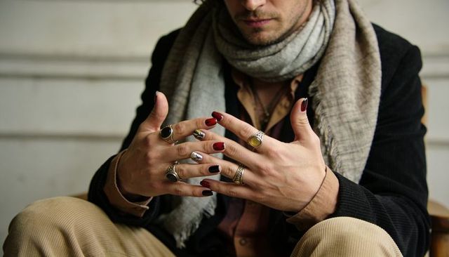 Displaying seated man showing ornate gemstone rings and painted nails wearing scarf