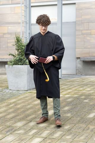 Young graduate wearing black gown holding mortarboard and burgundy diploma cover outdoors