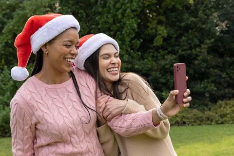 Multicultural friends wearing santa hats taking laughing holiday selfie in backyard