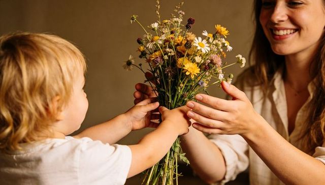 Toddler handing wildflower bouquet to smiling mother, tender warm family moment closeup