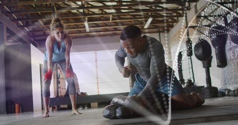 Kneeling boxer powering floor-bag punch while training partner watches in sunlit warehouse gym