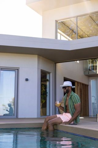 Man sitting on luxury modern pool deck wearing wide hat and sunglasses holding drink