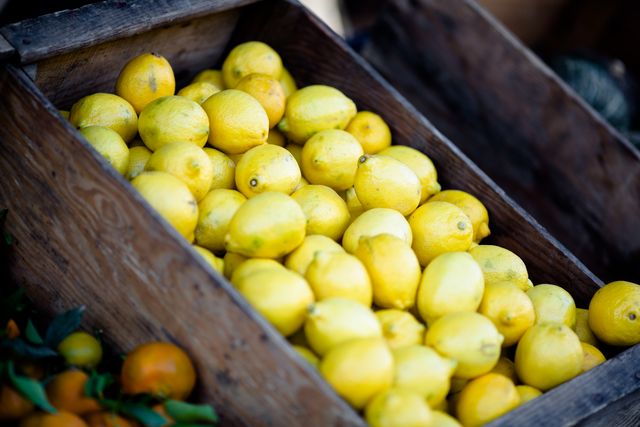 A pile of bright yellow lemons displayed in a rustic wooden crate, suggesting an outdoor market stall. The fresh lemons evoke a sense of natural, healthy eating and farm-to-table freshness. Use this image for themes related to organic food, health and wellness, farmers' markets, and rustic aesthetics in cooking blogs, health articles, and promotional material for natural products.