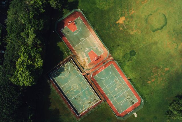 Aerial view captures three outdoor basketball courts surrounded by lush green park area. The uncluttered courts show wear and indicate regular use. Ideal for illustrating sports facilities, community parks, recreational spaces, outdoor activities, and healthy lifestyle concepts.