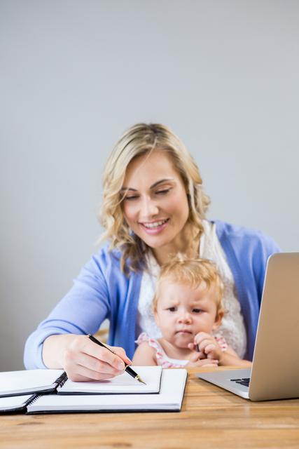 Mother holding baby girl while writing notes in personal organizer at home