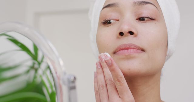 Young woman applying a skincare product to her face while looking in the mirror. Suitable for articles or promotions related to beauty, skincare routines, self-care tips, and cosmetic product advertisements.