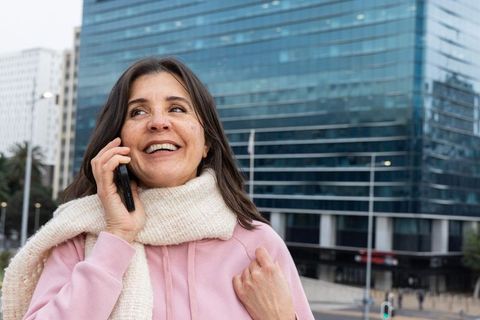 Smiling middle-aged woman talking on phone downtown wearing pink hoodie and cream scarf