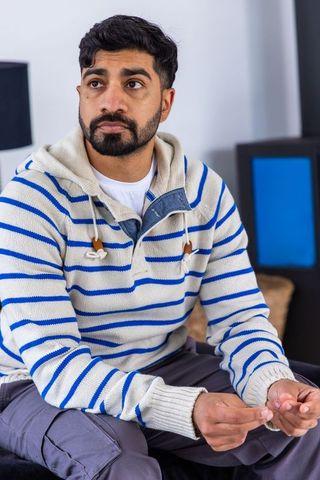 Contemplative asian man sitting on sofa wearing striped hoodie and denim shirt modern home portrait