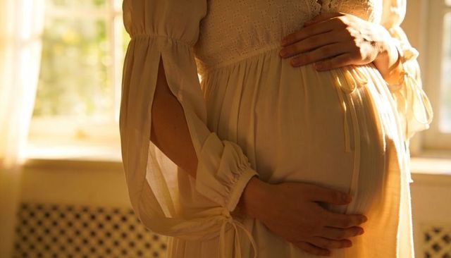 Sunlit maternity moment cradling pregnant belly in flowing cream gown with ribbon ties