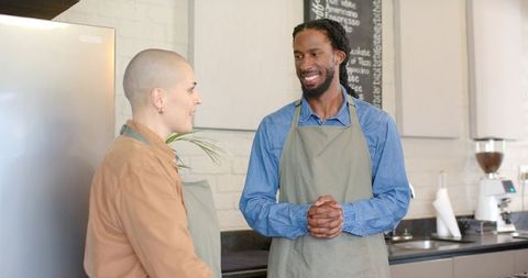 Baristas chatting and smiling behind cafe counter wearing aprons, teamwork and hospitality