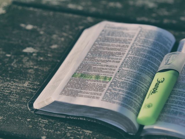 Open Bible lying on a rustic wooden table with a green highlighter highlighting text. Ideal for use in faith-based educational materials, religious studies promotions, spiritual blog posts, or articles about personal study practices. This image can also be used in advertisements for religious institutions, learning tools, or study aids.