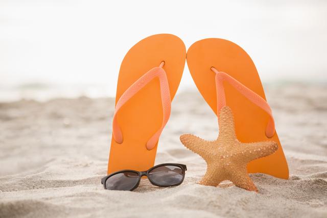 Orange flip flops and a starfish are placed on sandy beach with sunglasses nearby. Ideal for promoting summer vacations, beach holidays, travel destinations, and leisure activities. Perfect for use in travel brochures, summer-themed advertisements, and social media posts about beach trips.