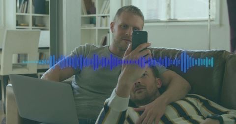 Same-sex couple listening to voice message on smartphone with laptop and audio waveform