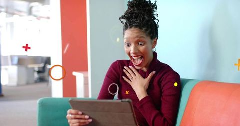 Excited woman reacting to tablet while sitting on colorful sofa in modern office lounge